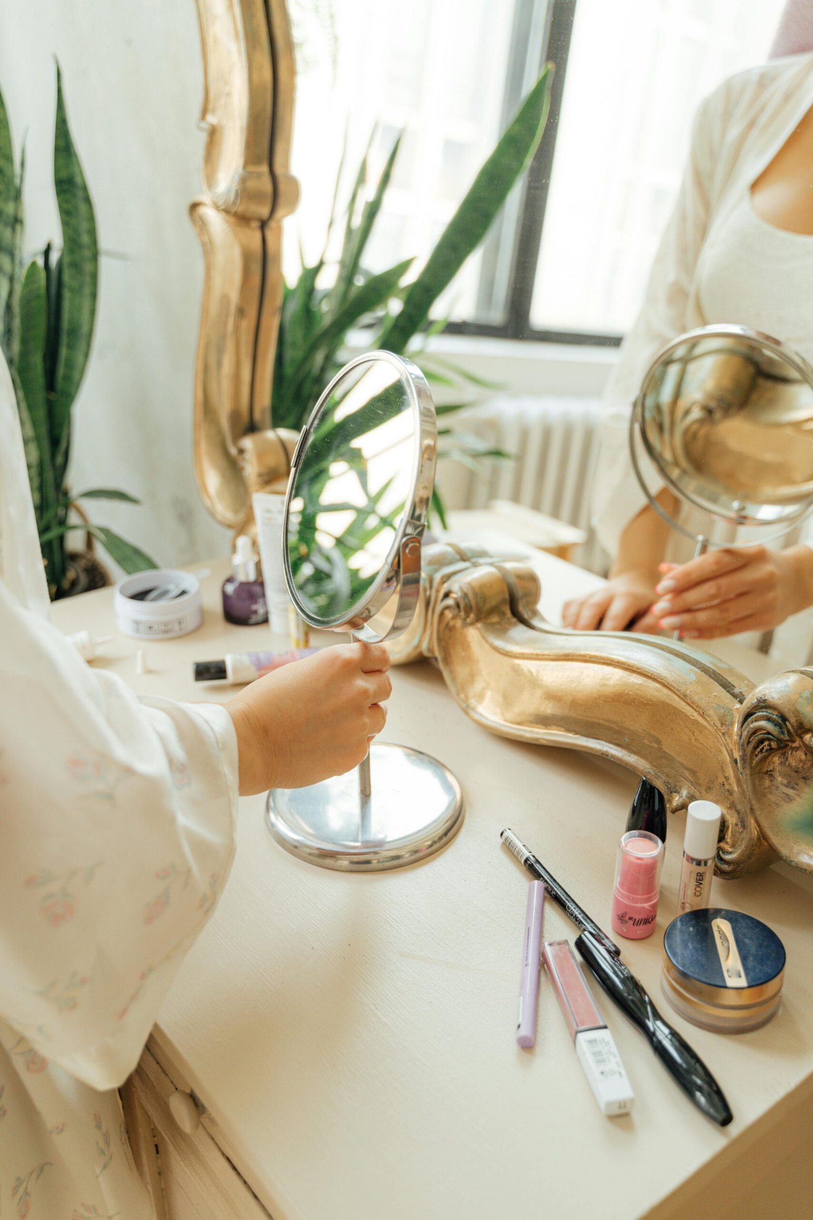 A woman's hands holding a mirror surrounded by cosmetics on an elegant vanity.